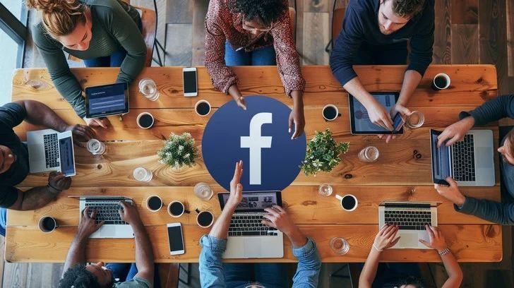 A group of office workers sitting around a wooden table with laptops and a big Facebook icon on the table 